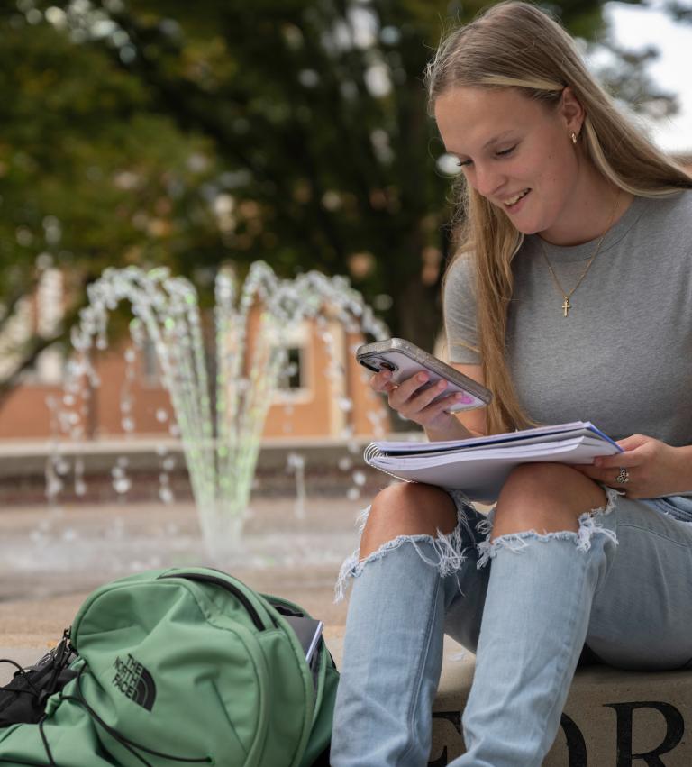 student sitting around the campus fountain smiling and looking at their phone.