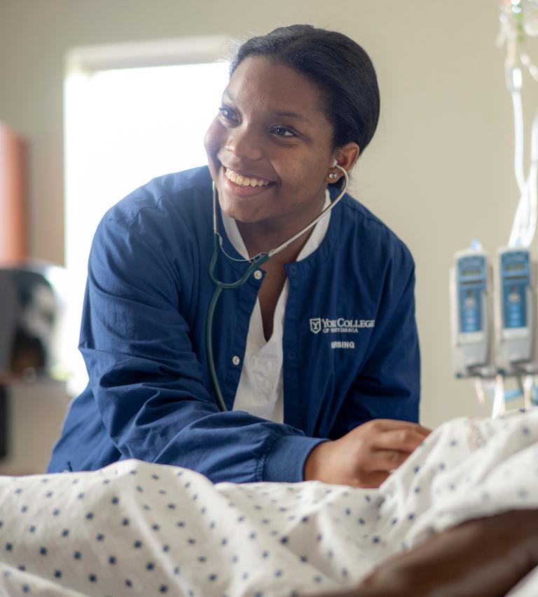 A nursing student smiles while listening to the heartbeat of a mannequin in the nursing simulation lab.