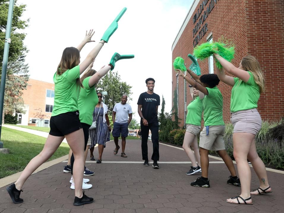 A group of cheerful student ambassadors dressed in green t-shirts cheer and wave green pom poms as a new student approaches.