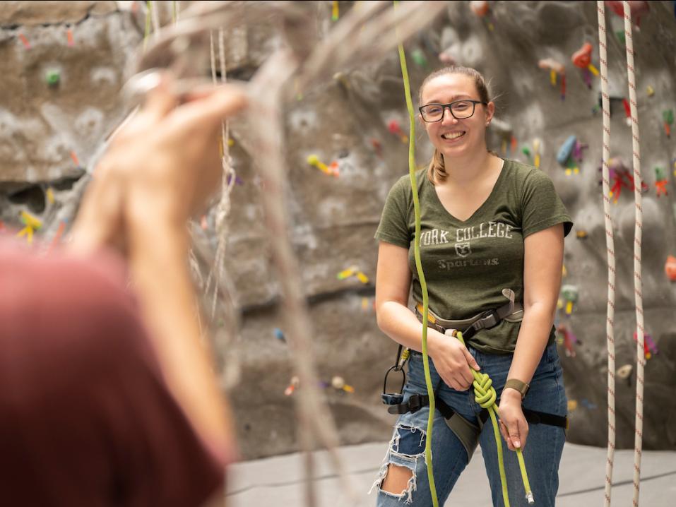 A student smiling while gearing up to climb the rock wall.
