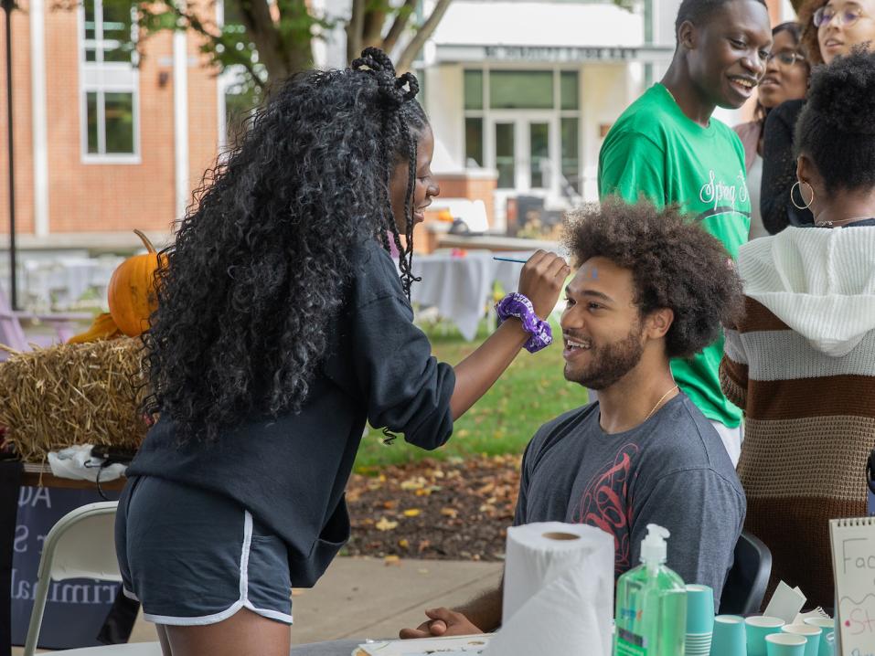 One student face paints another student during an outdoor involvement fair on campus.