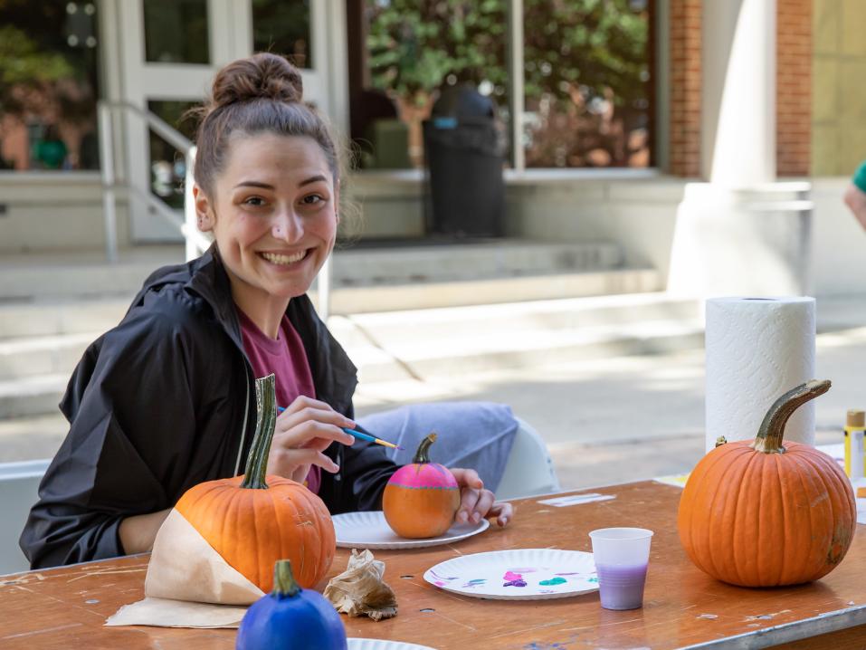A student paints a pumpkin at an outdoor activity fair on campus.