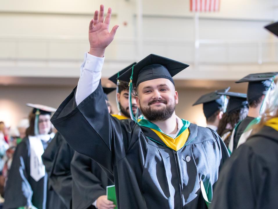 As students process into the commencement ceremony dressed in graduation regalia, one graduate waves and smiles broadly at the camera.
