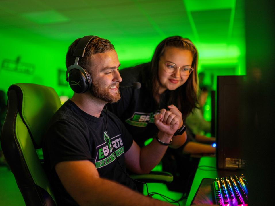 Two students celebrate a win as one plays a video game in the esports room. A green light is cast throughout the darkened room.