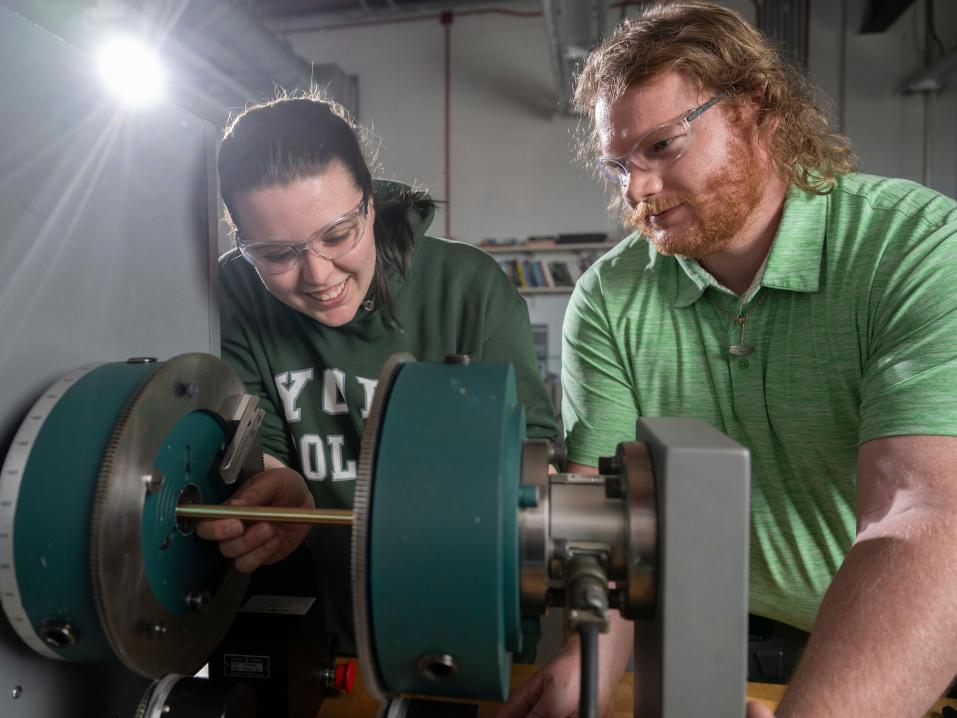 Two students smile as they wear safety goggles and examine machinery in the mechanical engineering lab.