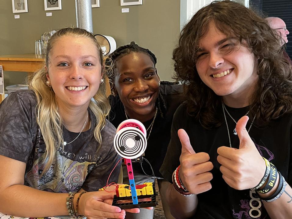 Three students sit together smiling at the camera with a lego creation