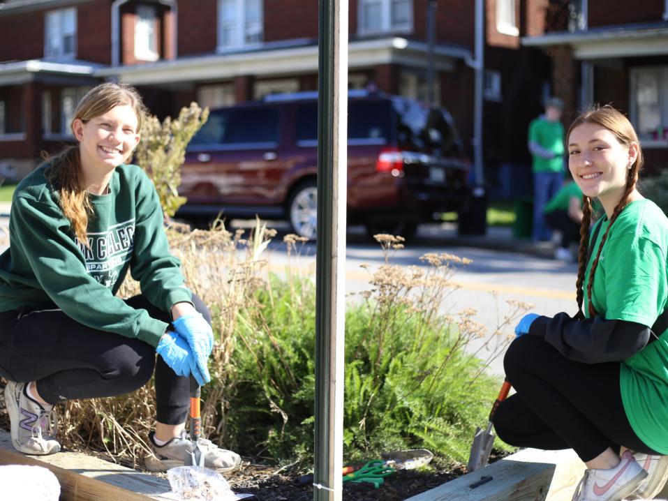 Two female students smile at the camera while kneeling next to a garden on Jackson Street.