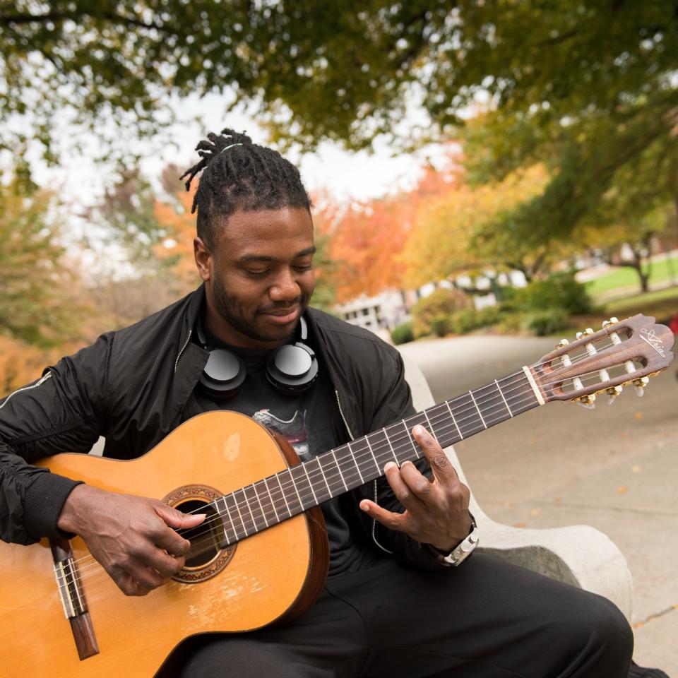 Student sits on bench and plays acoustic guitar