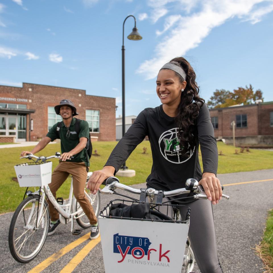 Two students ride bikes together on a campus pathway