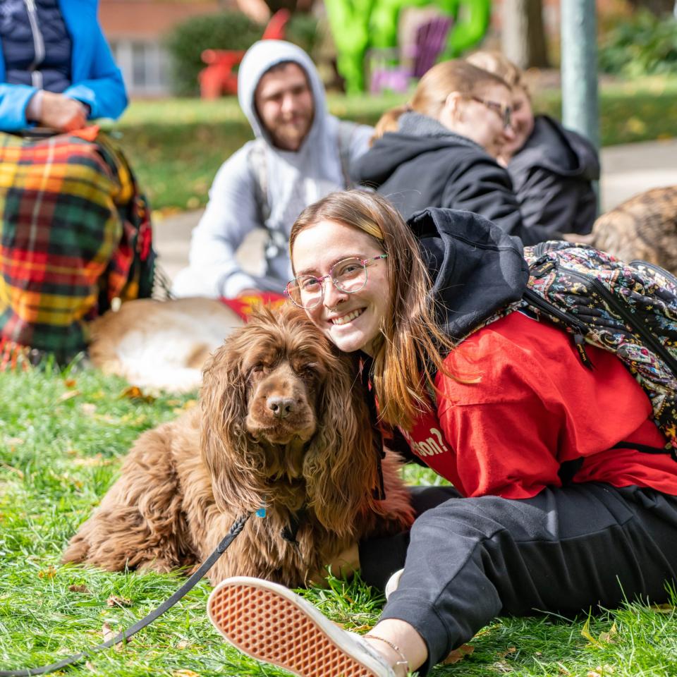 Students sit together on campus lawn, one is posing with a therapy dog