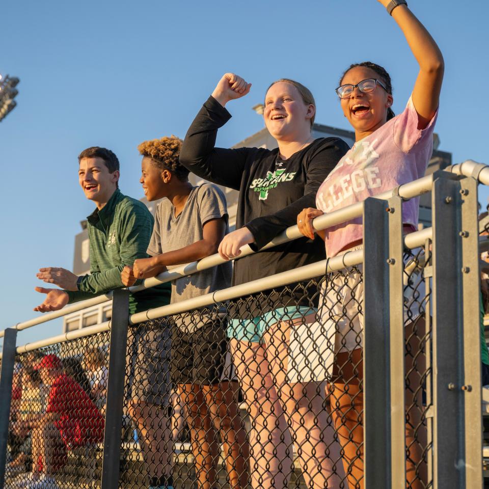 Four students cheer from the bleachers during a sunset lacrosse game.