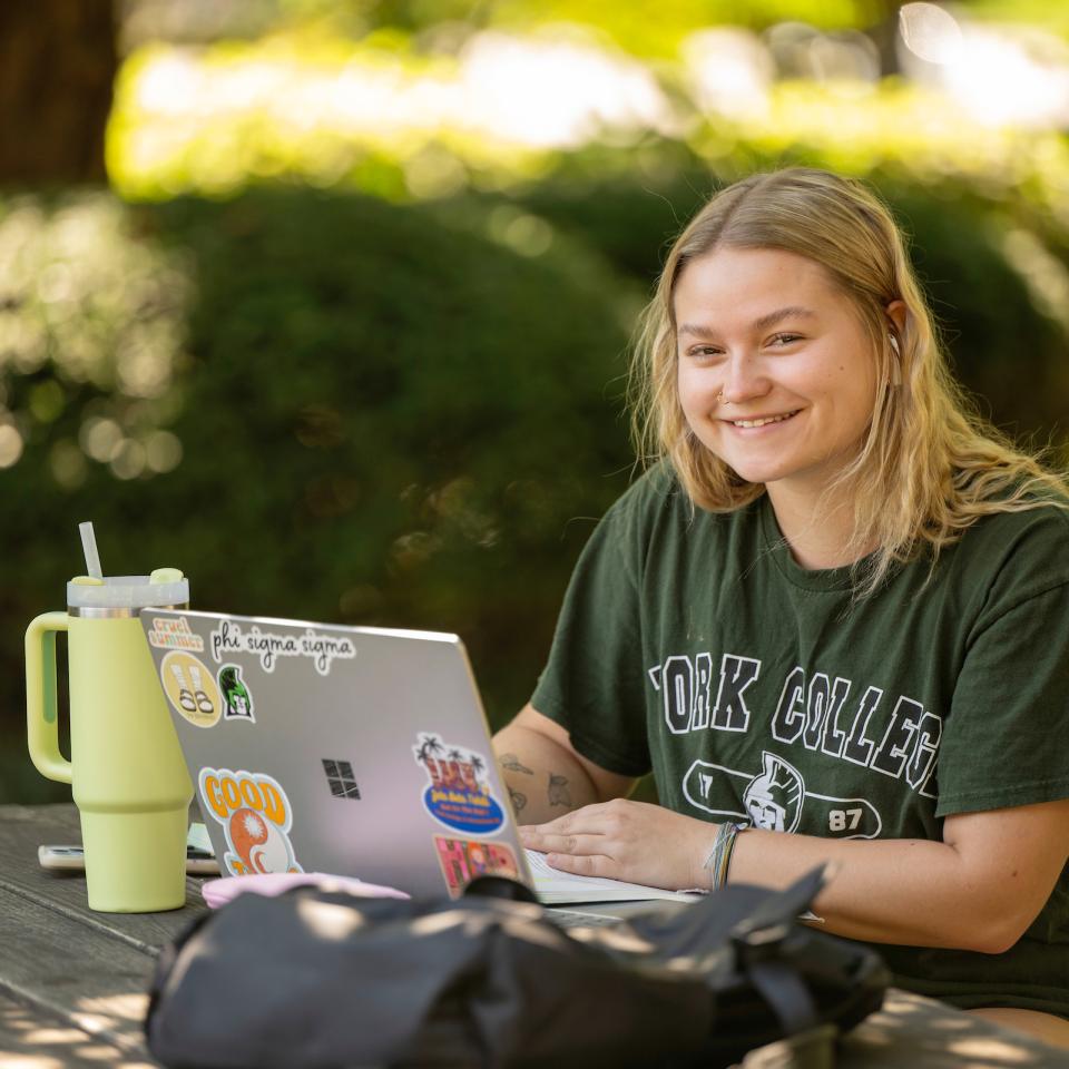 A student smiles from a picnic table on main campus as she works on her laptop.