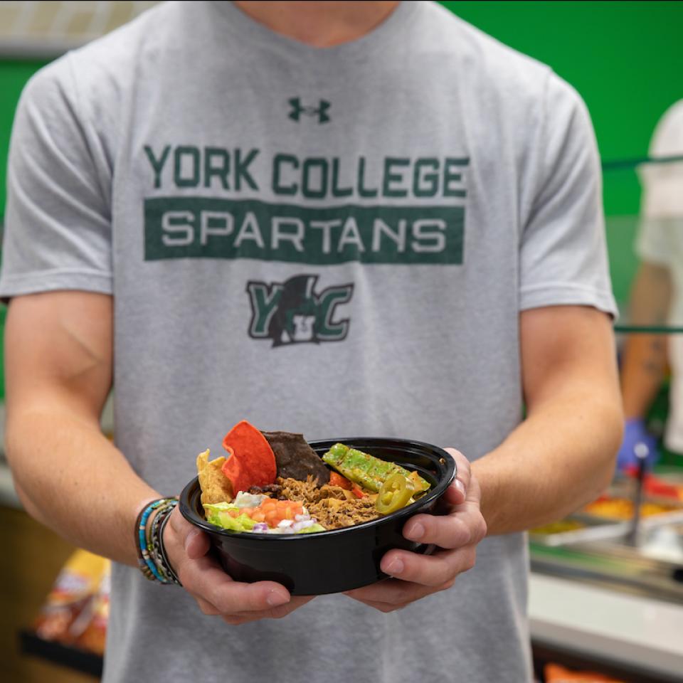 A student in a York College Spartans t-shirt holds a burrito bowl.