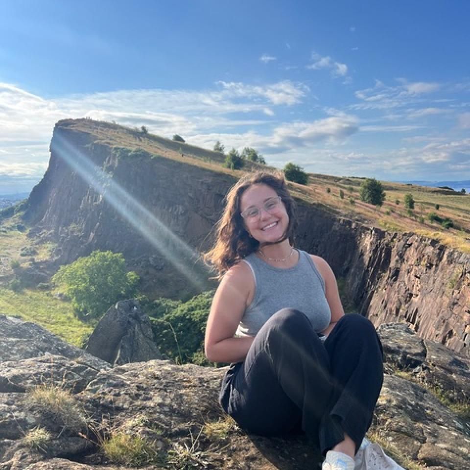 Gia Levis smiles while sitting on a rock outcropping in Edinburg, Scottland on a sunny day.