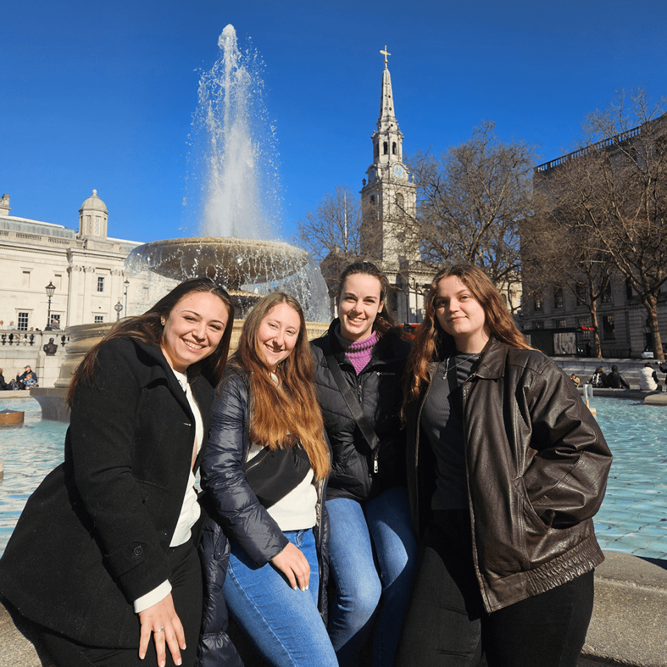 Four students from the International Sport and Entertainment course pose in front of a fountain with a clear blue sky and architectural spire in the background.