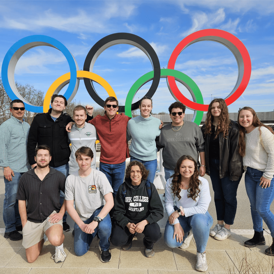 A group of individuals from the International Sport and Entertainment course at York College pose in front of Olympic rings.