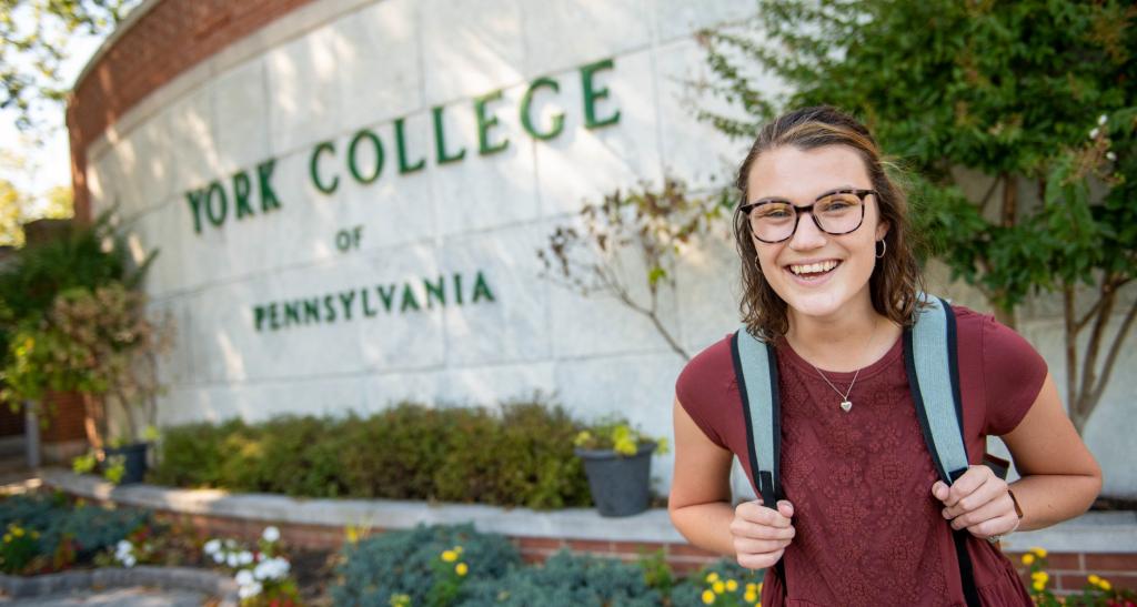 Student in red shirt with backpack smiles in front of York College entrance sign