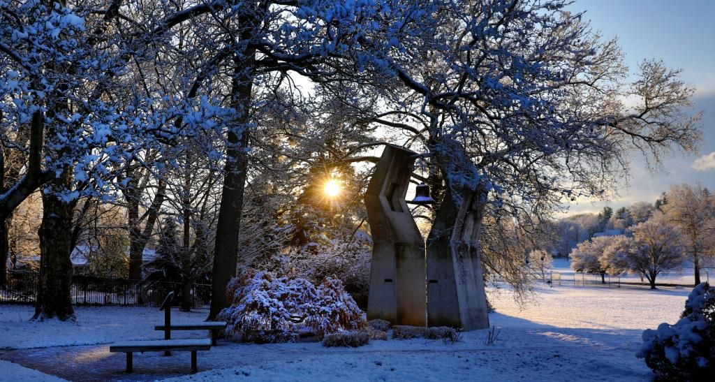 A stone monument that resembles prayer hands with a brass bell in-between them. Winter setting with snow on the ground and trees.