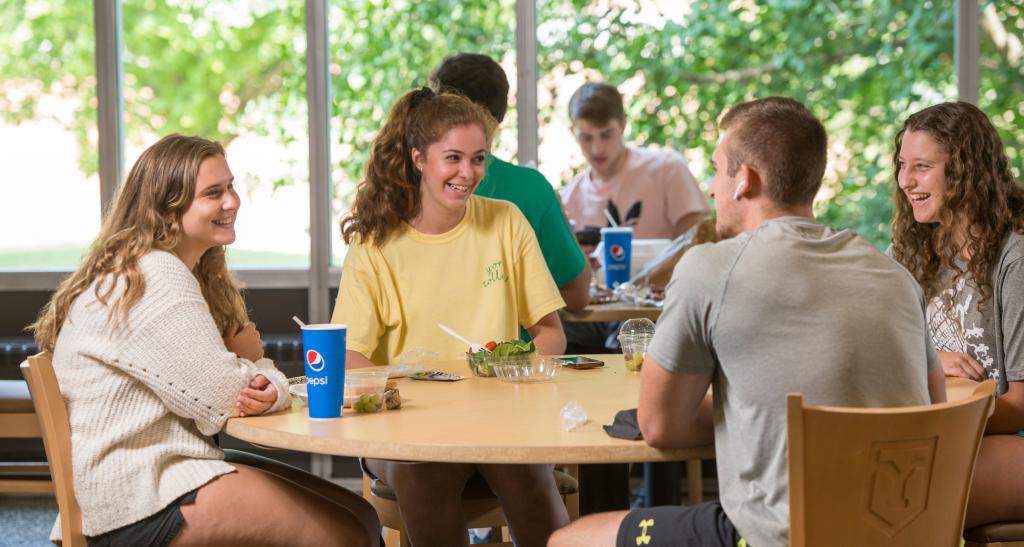 Four students laugh over lunch at a round table in the dining hall.