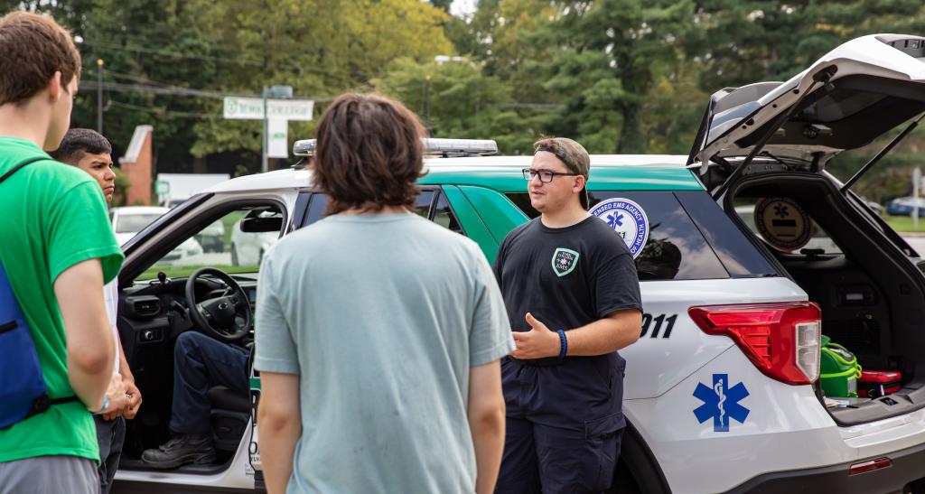 A student in an EMS t-shirt stands in front of an EMS vehicle with an open trunk, speaking to a group of students.