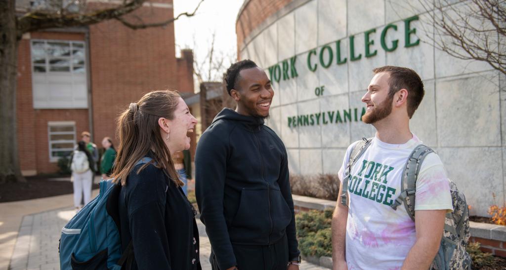 Three students laughing and talking by the York College of Pennsylvania sign.