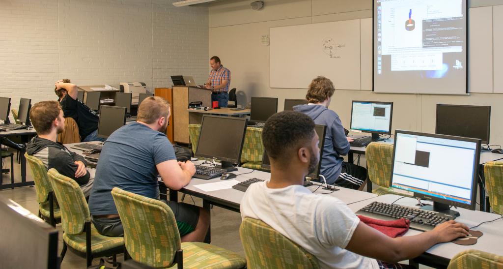 A class of students work in the computer lab while a professor lectures at a podium at the front of the room.