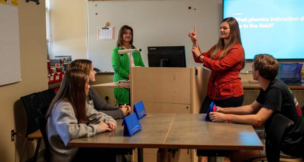 A teacher presents at the front of a classroom; a projection screen is visible behind her.