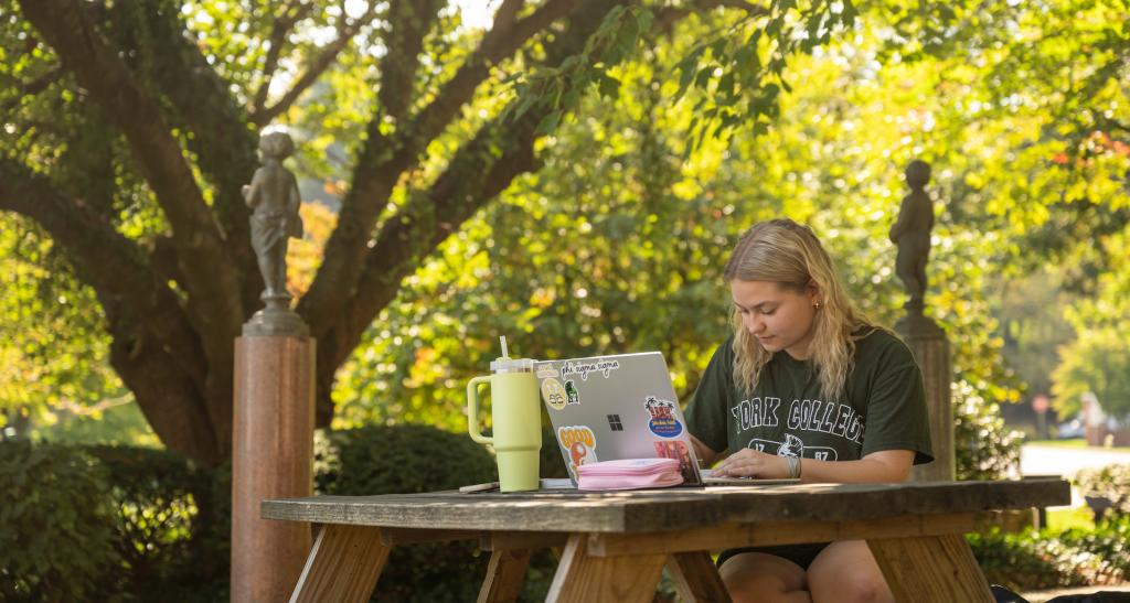 A student studies with her notes and laptop at an outdoor picnic table on main campus.