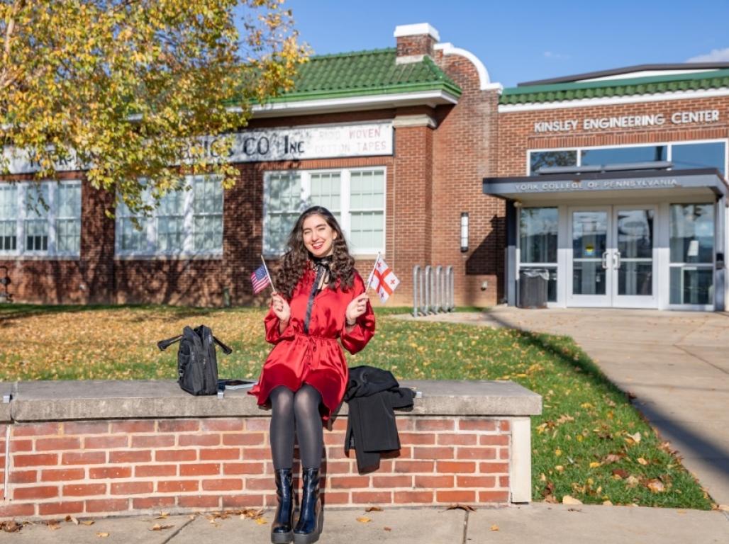 Baia Grdzelishvili sits on low wall in front of the Kinsley Engineering Center, waving a United States and country of Georgia flags.