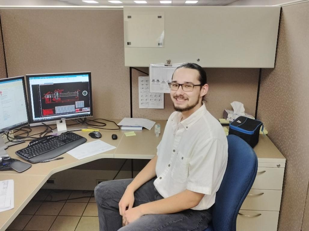 Brandon Goromaru sits at a desk with multiple computer monitors and documents.