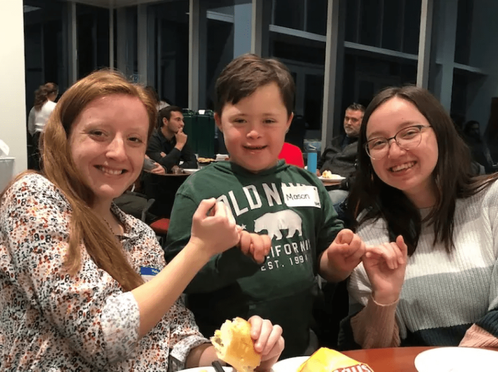 Two education majors sit at a dining table, making pinky promises and smiling with a child with Down Syndrome.