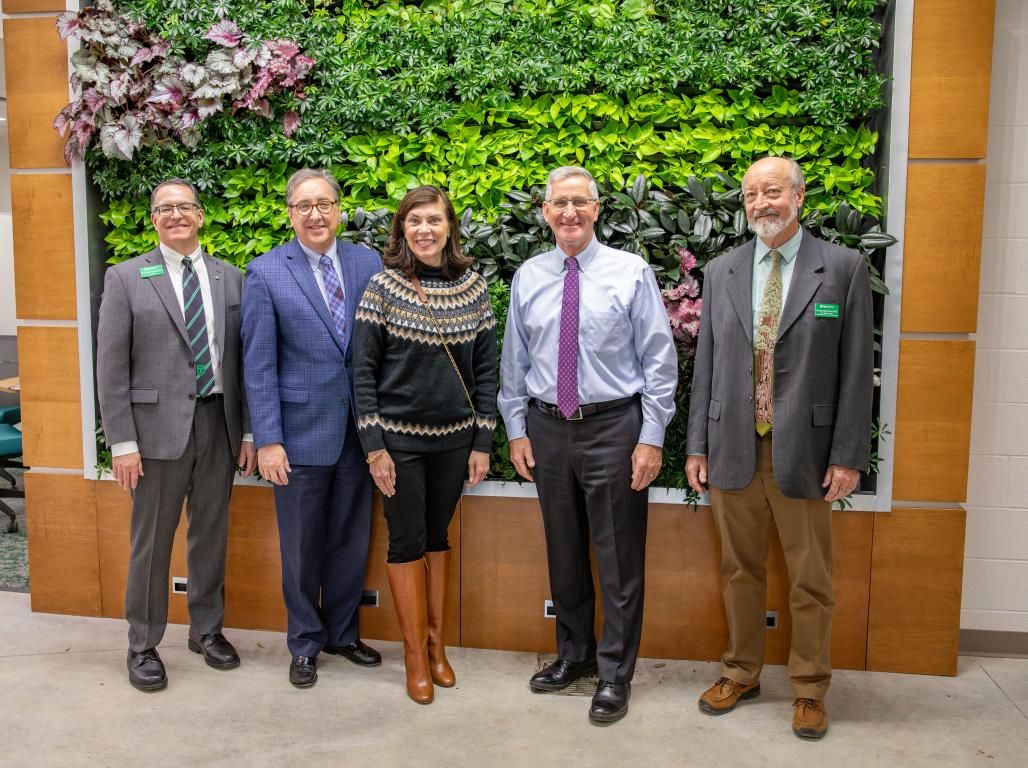 A group of five including visiting state government representatives, President Thomas Burns, and faculty pose in front of a wall of plants in the Appell Horticulture Center. 