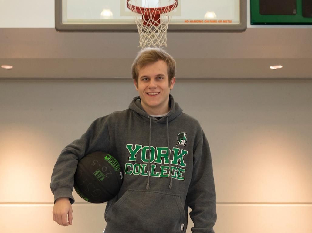 Jacob Carlson stands in a gymnasium beneath a basketball hoop, holding a ball under one arm and wearing a York College sweatshirt.