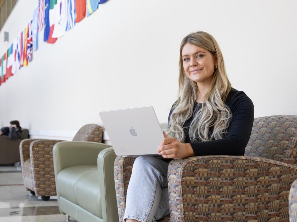 Marianna Lally on her laptop smiling at the camera, sitting in a hallway decorated with international flags.