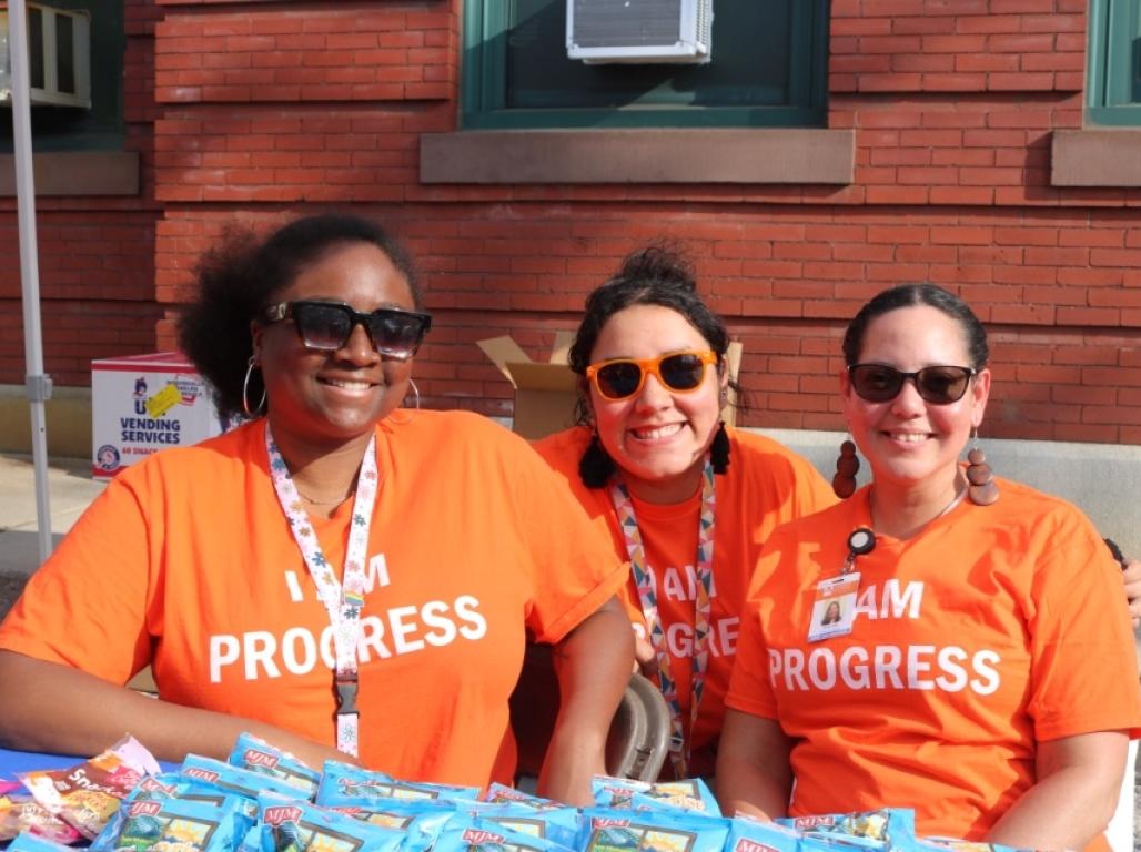 Melissa St. Cloud and her team wearing orange t-shirts printed with the words "I Am Progress" sit at a table of healthy snacks.
