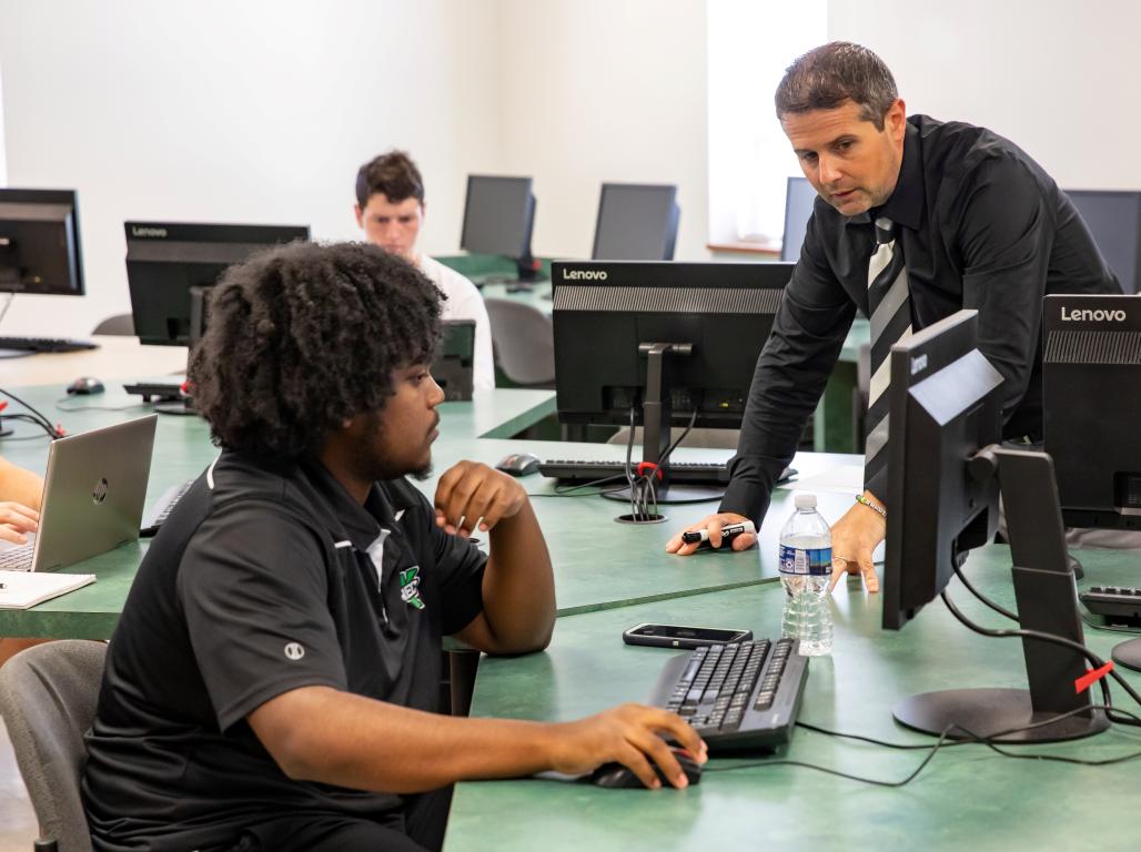 Mike Mudrick leans on a table to assist a student working on a computer.