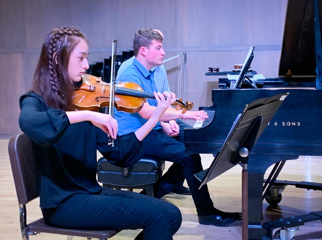 Students play orchestral instruments on stage.