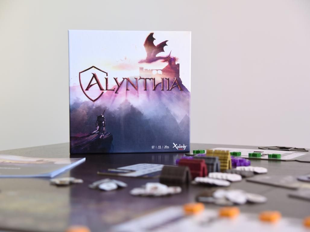 A mock-up shows the box cover of the board game Alynthia, featuring a dragon and fantastical artwork. Game cards and pieces are scattered on the table in front of the box.