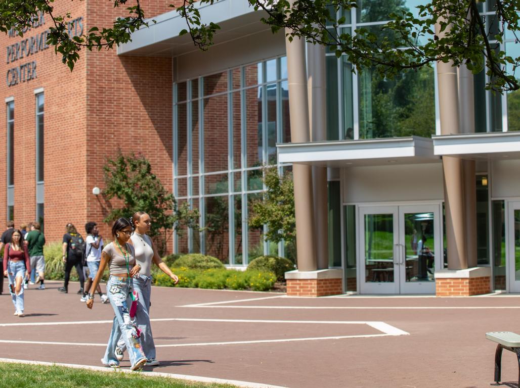 Students walk across campus on a sunny day, passing in front of the multi-story glass windows of the Performing Arts Center.