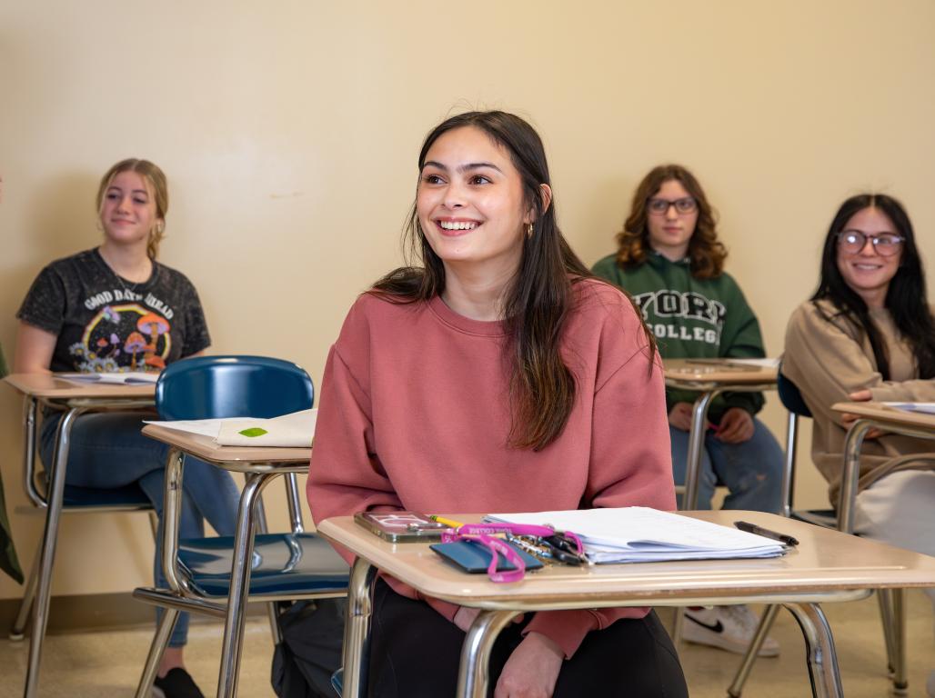 A York College student smiles as she sits in a classroom desk/chair combo. Classmates are visible behind her, also seated in desk/chairs.