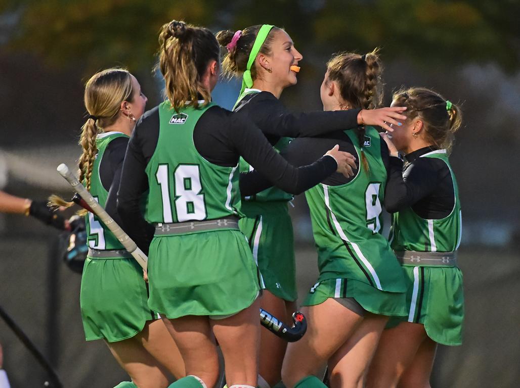 Smiling York College field hockey players, wearing green uniforms, circle around in celebration.