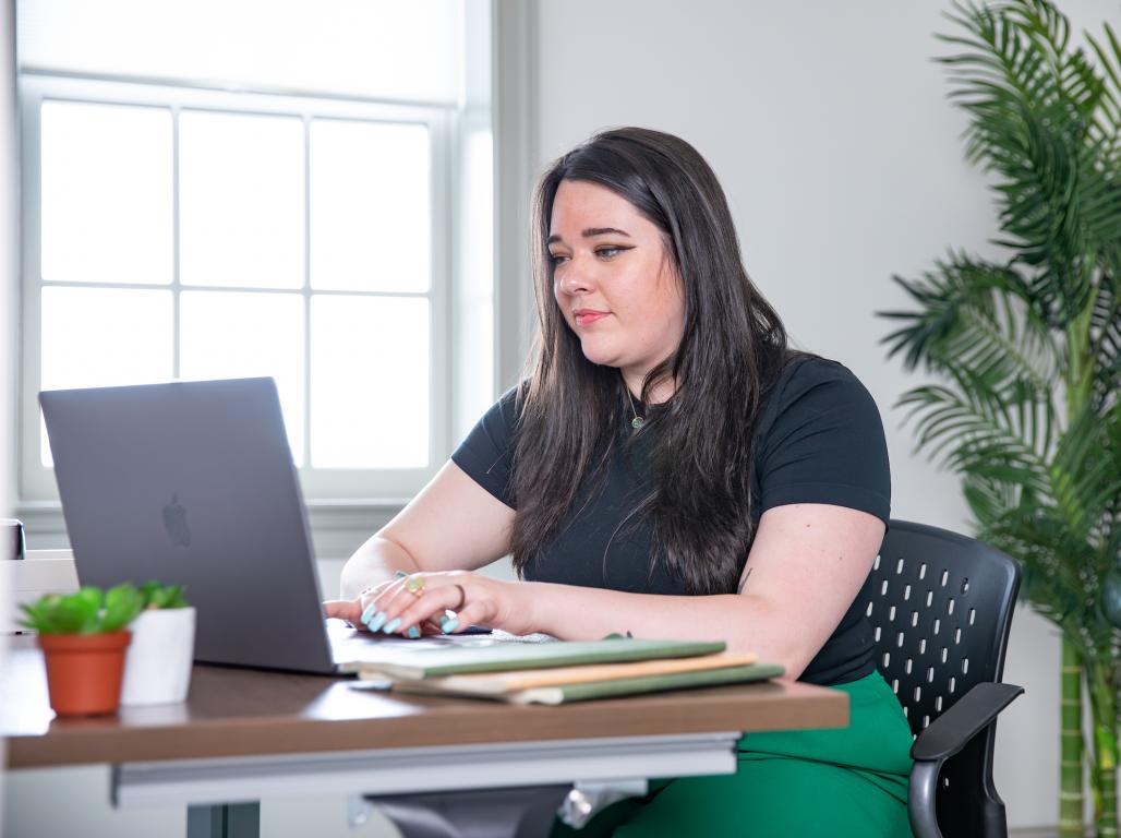 A female professional wearing a black shirt and green pants is sitting at a desk working on a laptop.