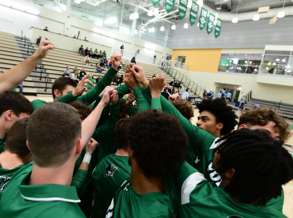 The Men's Basketball team at York College gathers in a huddle on the court, fists raised in the air.