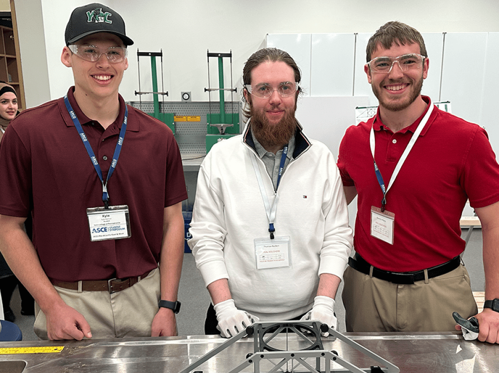 Three individuals wearing safety glasses and lanyards smile at the camera in an engineering lab with a 3D printed bridge in the foreground. 