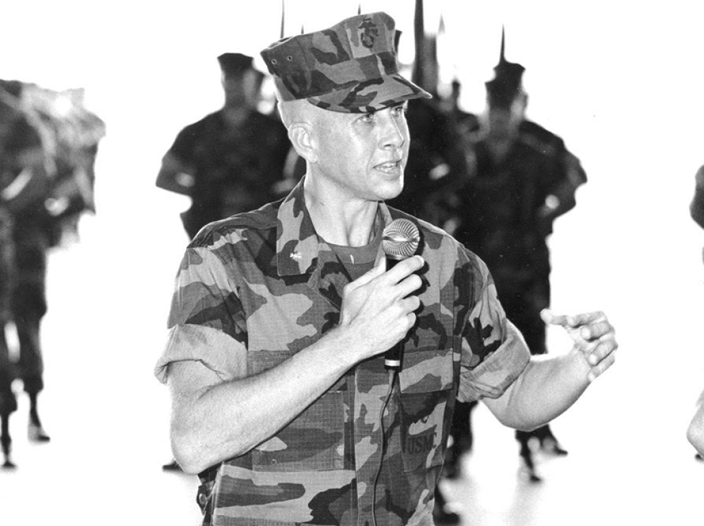 An individual wearing military fatigues speaks into a microphone with soldiers standing out of focus in the background.