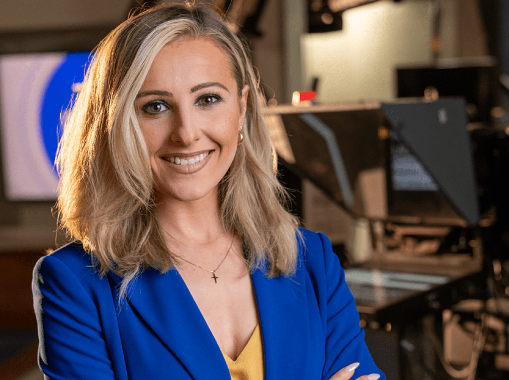 Person wearing a blue blazer smiles at the camera in a TV studio setting. 