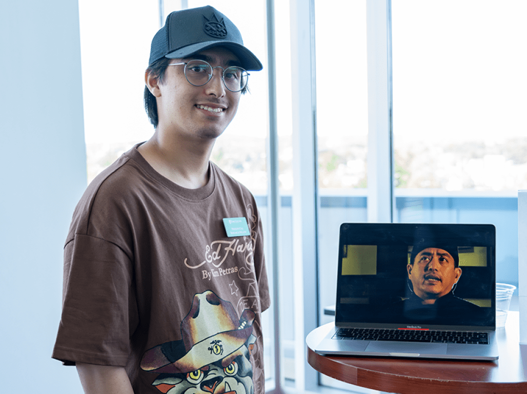 Student smiles at the camera while showcasing his short film on a laptop.
