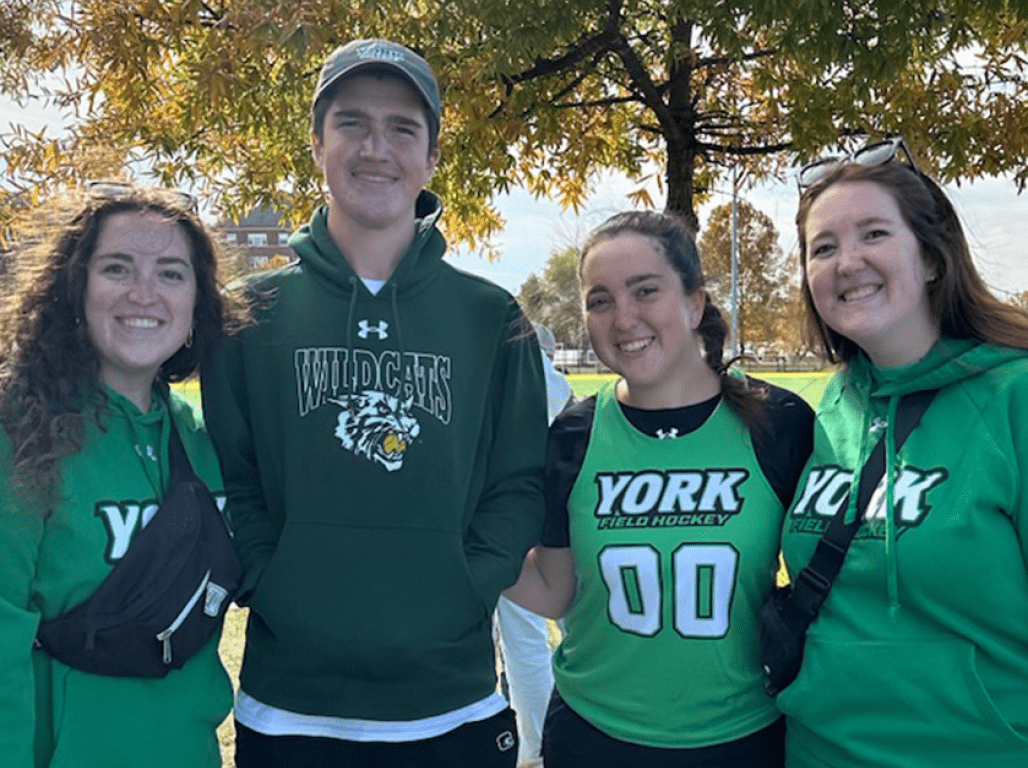 Four people smiling at the camera wearing green York College shirts at a sporting event on a fall day.