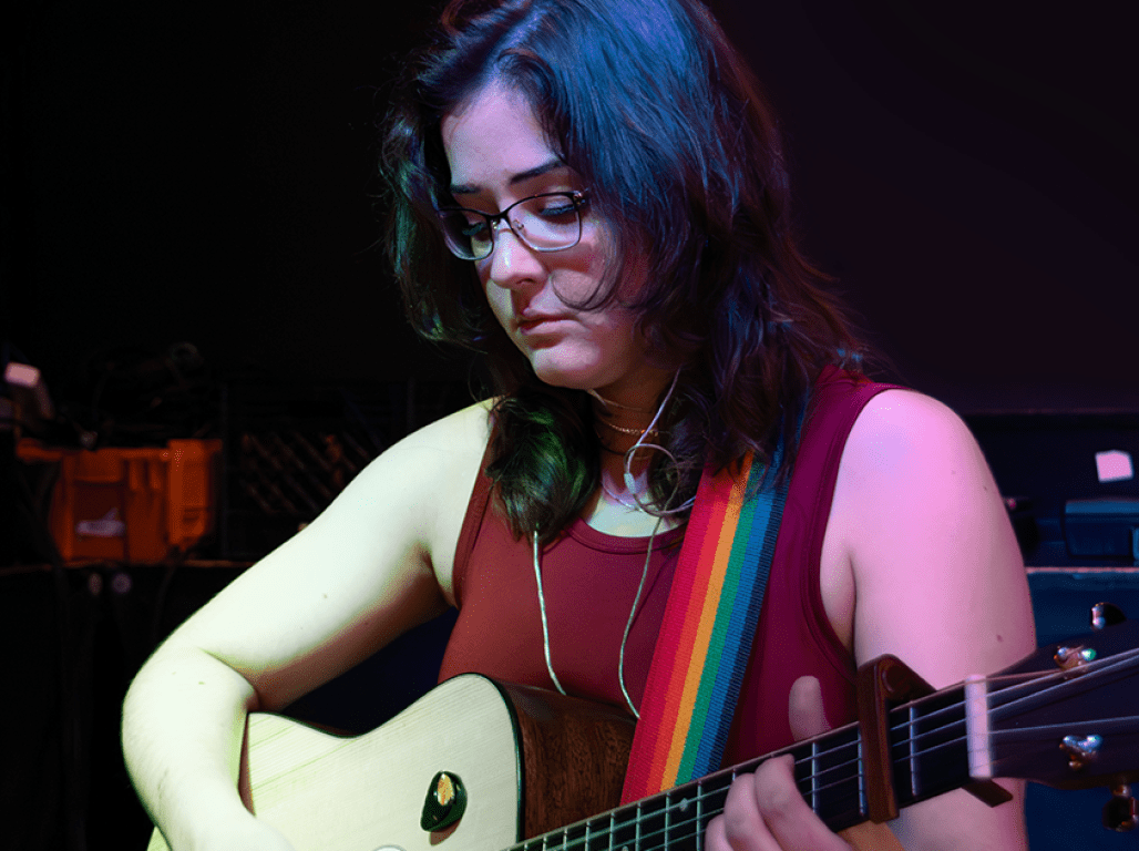 Person playing a guitar with a rainbow guitar strap under stage lighting.