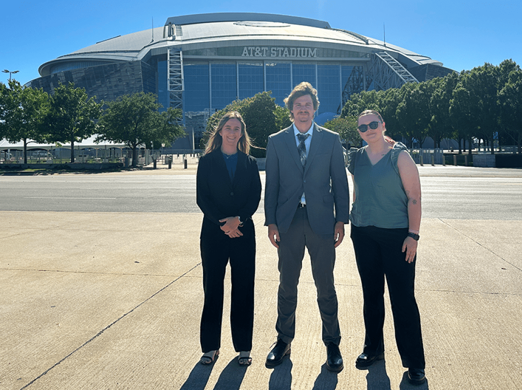 Three people smile at the camera in front of the AT&T stadium.